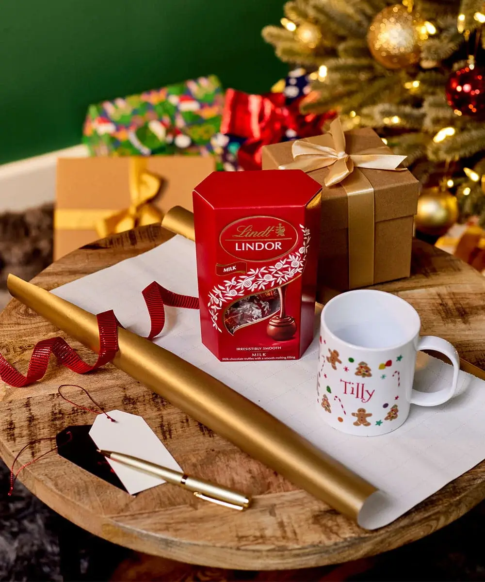Christmas gift wrapping scene with chocolates, a personalised festive mug, wrapped presents, gift tags, and wrapping paper beside a decorated Christmas tree. Christmas gift wrapping scene with chocolates, a personalised festive mug, wrapped presents, gift tags, and wrapping paper beside a decorated Christmas tree.